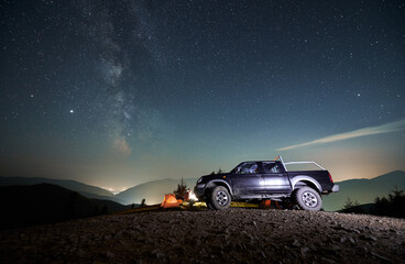 Obraz na płótnie Canvas Black SUV car parked on dirt mountain road near tourist camp with orange tent under night starry sky, against the backdrop of silhouettes of mountain peaks and city lights in the distance.