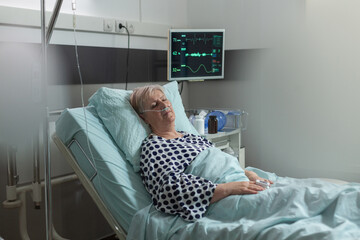 Senior woman patient in hospital room getting medicine through intravenous line from iv drip bag and breathing with help from oxygen mask laying in bed. Oxymeter attached on finger monitoring bpm.