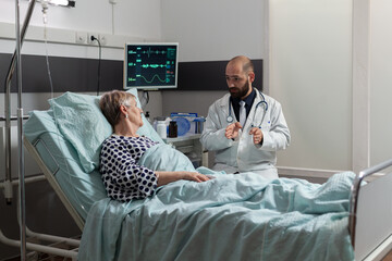 Fototapeta premium Doctor sitting next so sick unwell senior patient laying in bed during friendly discussion about treatment. Breathing with help from oxygen tube and getting medication through iv drip bag.