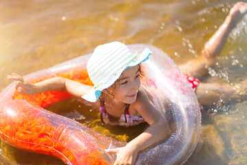 Girl in a hat swimming in the river with a transparent inflatable circle in the shape of a heart with orange feathers inside.The sea with a sandy bottom. Beach holidays, swimming, tanning, sunscreens.