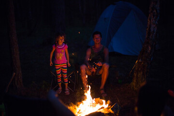 Dad and daughter sit at night by the fire in the open air in the summer in nature. Family camping trip, gatherings around the campfire. Father's Day, barbecue. Camping lantern and tent