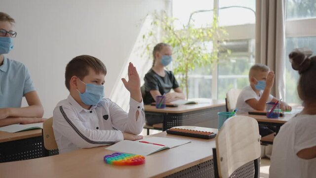 Raise your hand to answer the teacher's question. Multi-ethnic group of children with face masks at school during COVID-19 pandemic.