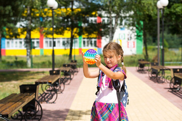 Little girl with a backpack and in a school uniform in the school yard plays pop it toy. Back to school, September 1. The pupil relaxes after lessons. Primary education, elementary class for student