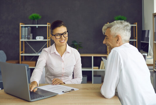 Smiling retirement plan advisor, bank employee or insurance company worker meeting senior client. Young woman sitting at office table with laptop showing good pension plan details to happy older man - Powered by Adobe