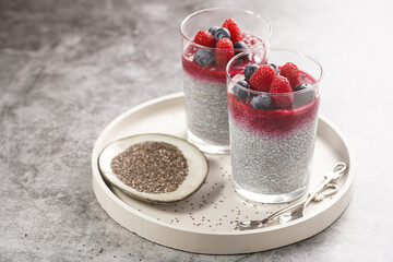 Two glasses with chia seeds pudding, raspberry jam and fresh raspberries and blueberries on a concrete tray on grey surface