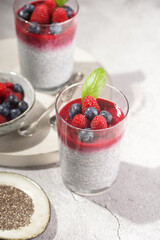 Two glasses with chia seeds pudding, raspberry jam and fresh raspberries and blueberries on a concrete tray on grey surface, a close up