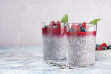 Two glasses with chia seeds pudding, raspberry jam and fresh raspberries and blueberries on a concrete tray on grey surface