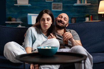 Relaxed couple sitting on couch watching movie on television, drinking beer playing popcorn games enjoying time together at home leisure, happiness and married people concept.