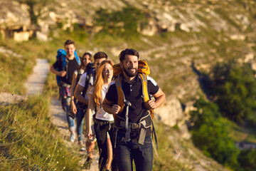 Group of young smiling friends hikers walking along mountain valley in row together on summer vacations on sunny clear day, selective focus. Hiking, traveling with backpack, vacations concept