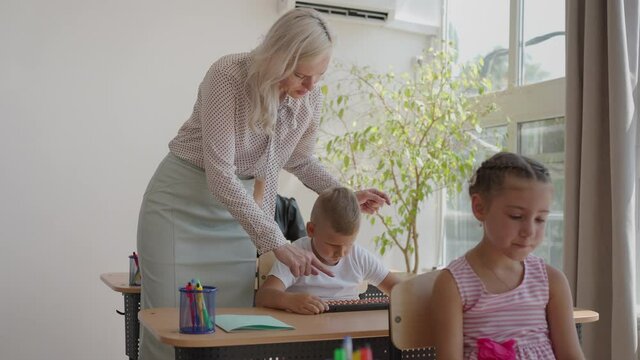 In Elementary School Class: Enthusiastic Teacher walks between Rows of Bright Diverse Children, Explains Lesson. Group of Smart Multiethnic Kids Learning New Stuff. Side View Tracking Shot