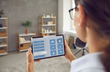 Woman controlling Smart Home with touchscreen tablet. Young lady using mobile interface with wi fi, faucet etc icons to monitor house system. People and technology concept. Close up view over shoulder