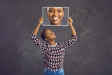 Happy pretty young black woman with short hair holding photo of her face with clear skin and...