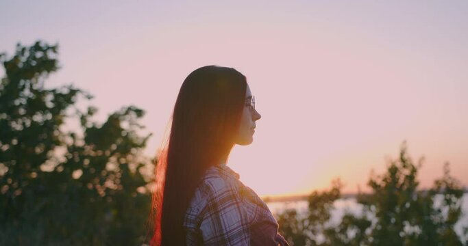 A young red-haired girl on the background of the sunset is illuminated by the rays of the sun, looks thoughtfully into the distance