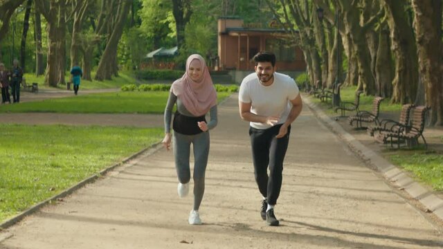 Positive young people in sport clothes running together at morning park. Handsome muslim husband and charming wife in hijab training actively outdoors. Happy arabian couple in active wear jogging