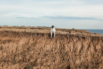 a man with a beard sits on the edge of a cliff with his legs dangling down in summer
