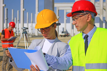 Female architect with clipboard and construction engineer in hardhats talking about the project on construction site, behind them construction worker with measuring device, teamwork