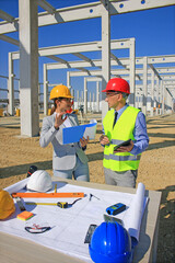 Female architect with clipboard and construction engineer with tablet in hardhats talking about the project on construction site, behind them construction worker with measuring device, teamwork