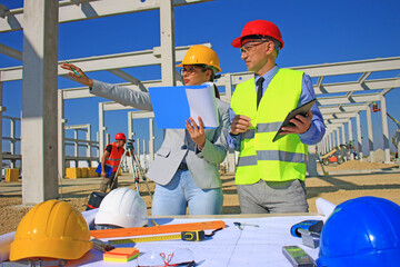 Female architect with clipboard and construction engineer with tablet in hardhats talking about the project on construction site, behind them construction worker with measuring device, teamwork