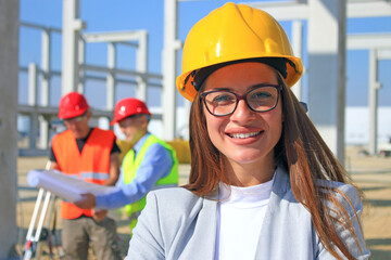 Happy beautiful female architect on construction site. She is smiling and satisfied with her job, behind her construction engineer and worker talking about the project, teamwork positive emotions