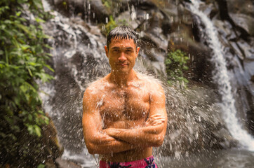 A handsome fit brunet man smiling as he is standing under shower of a river waterfall in the mountains. A male traveler on a hiking trip in tropical jungle under defocused cold streams