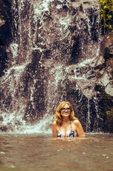 A beautiful blonde woman in her 50-s swimming in a river under waterfall in a tropical jungle in summer.