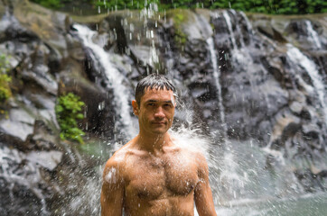 A happy strong male person under stream of cold waterfall in the mountains. Cold water training for a muscular athlete. Willpower and health, stamina and energy for sportsman in wild nature.