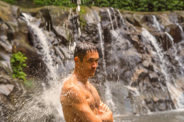 Fototapeta na wymiar A tired athlete taking shower under waterfall after training. Sportsman with muscular body standing under water streams on a mountain river.
