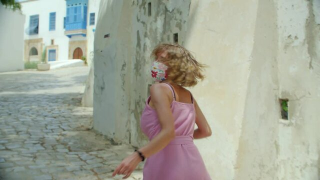 a tourist woman in a protective mask in the old city calls for her-follow me and runs ahead in her pink dress, in slow motion. a positive view of the new reality after the pandemic