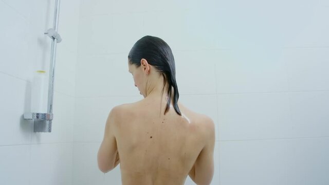 Back view of naked caucasian woman with dark hair taking shower at bright bathroom. Hygienic rituals for skin and body. Morning routine of charming lady.