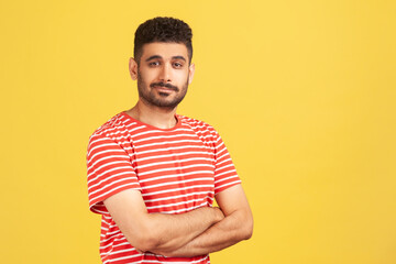 Portrait of serious bearded man in red striped t-shirt standing crossing hands on chest and looking at camera with confidence. Indoor studio shot isolated on yellow background