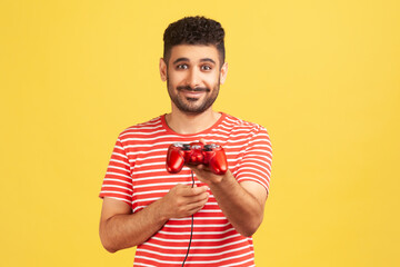 Generous bearded man in striped t-shirt holding in hand gaming joystick, suggesting you to play video game with him, virtual reality simulator. Indoor studio shot isolated on yellow background