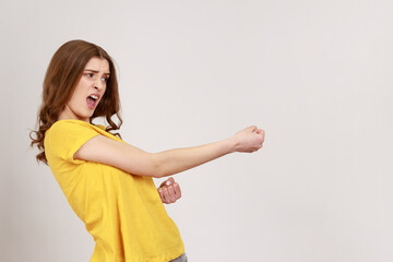 Side view portrait of teenager girl with brown wavy hair in casual style yellow T-shirt standing and doing pulling gesture of something. Indoor studio shot isolated on gray background.