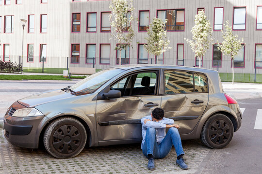 Man in despair wearing casual clothing sitting on ground with tilted head near broken car, damaged automobile with scratches and dents on doors, being sad of smashed vehicle. Outdoor shot.
