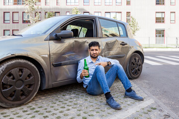 Upset bearded man wearing blue shirt and jeans sitting near smashed car and pointing at beer...