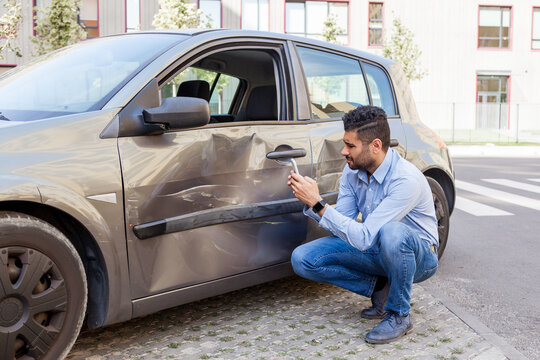 Portrait of brunette man wearing jeans and blue shirt not cope with driving, damaged his car door, dents and scratches on auto body, making photos of damaging for insurance inspection. Outdoor shot