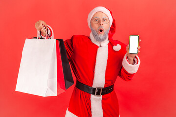 Surprised elderly man wearing santa claus costume holding shopping bags and phone with empty display, shocked by Christmas discounts, buying online. Indoor studio shot isolated on red background.