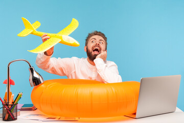 Dreamy man office worker playing with toy plane sitting at workplace with rubber lifebuoy,...