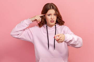 Portrait of teenage girl in hoodie showing stupid gesture with finger near head and blaming fool...