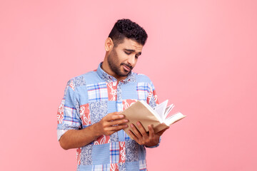 Young adult bearded man wearing blue casual style shirt standing with book in hands, reading boring novel with uninteresting plot. Indoor studio shirt isolated on pink background.