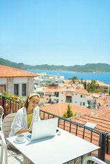 Pretty boho style girl sitting on terrace during summer vacation on Turkish resort, using laptop computer for chatting with friends in social network, reading good news