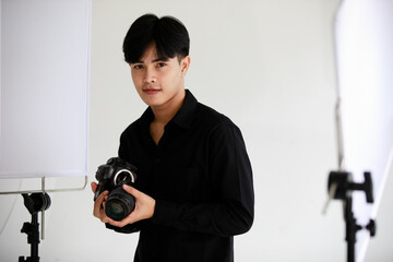 Asian young handsome successful male photographer wears black long sleeve shirt hold DSLR camera in hand while taking lens off from body in indoor photography studio full of lights and reflectors