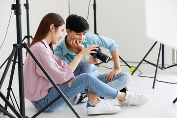 Fototapeta na wymiar Asian young friendly pretty success female photographer sit smile hold and point dslr camera screen in hands try to check choose best image and talk with handsome teen male model in photo studio