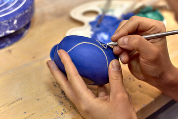 Hands of a ceramist who cuts a drawing with a knife on a painted blue cup in a workshop with a palette on the background