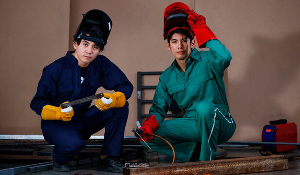 young smart Asian sitting and posing to a camera for photographing in a factory. They are wearing a safety outfit for factory working with a mechanic jumpsuit, glove, boots, and welding helmet