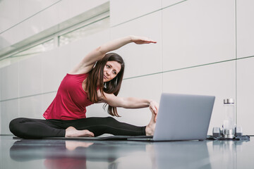 Young athletic fitness woman watching online leg stretching training video on yoga mat infront of a laptop at home studio indoor with panoramic window on reflective modern background 