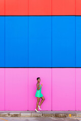 Stylish young black woman with electric green dress and white heels walking in front of a multicolored background and making V sign