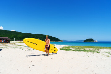 A man stands on a sandy shore and holds a large yellow paddle board.