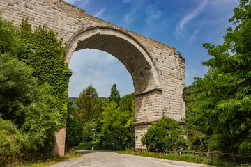 Fototapeta na wymiar The ruins of the Roman arch bridge of Augustus, in Narni, Terni, Umbria. The remains of the bridge over the Nera river. The big and ancient stone arch, against the blue sky. Trees and dense vegetation