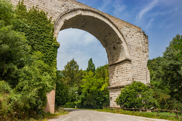 Fototapeta na wymiar The ruins of the Roman arch bridge of Augustus, in Narni, Terni, Umbria. The remains of the bridge over the Nera river. The big and ancient stone arch, against the blue sky. Trees and dense vegetation