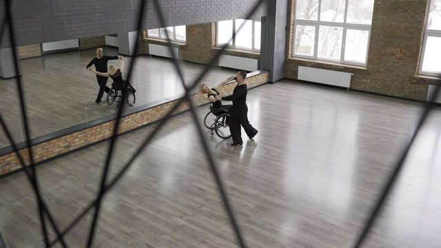 Top view of graceful couple, disabled female on wheelchair and her partner dancing holding hands in ballroom. Elegant pair of middle aged artists performing touching dance whirling hand in hand